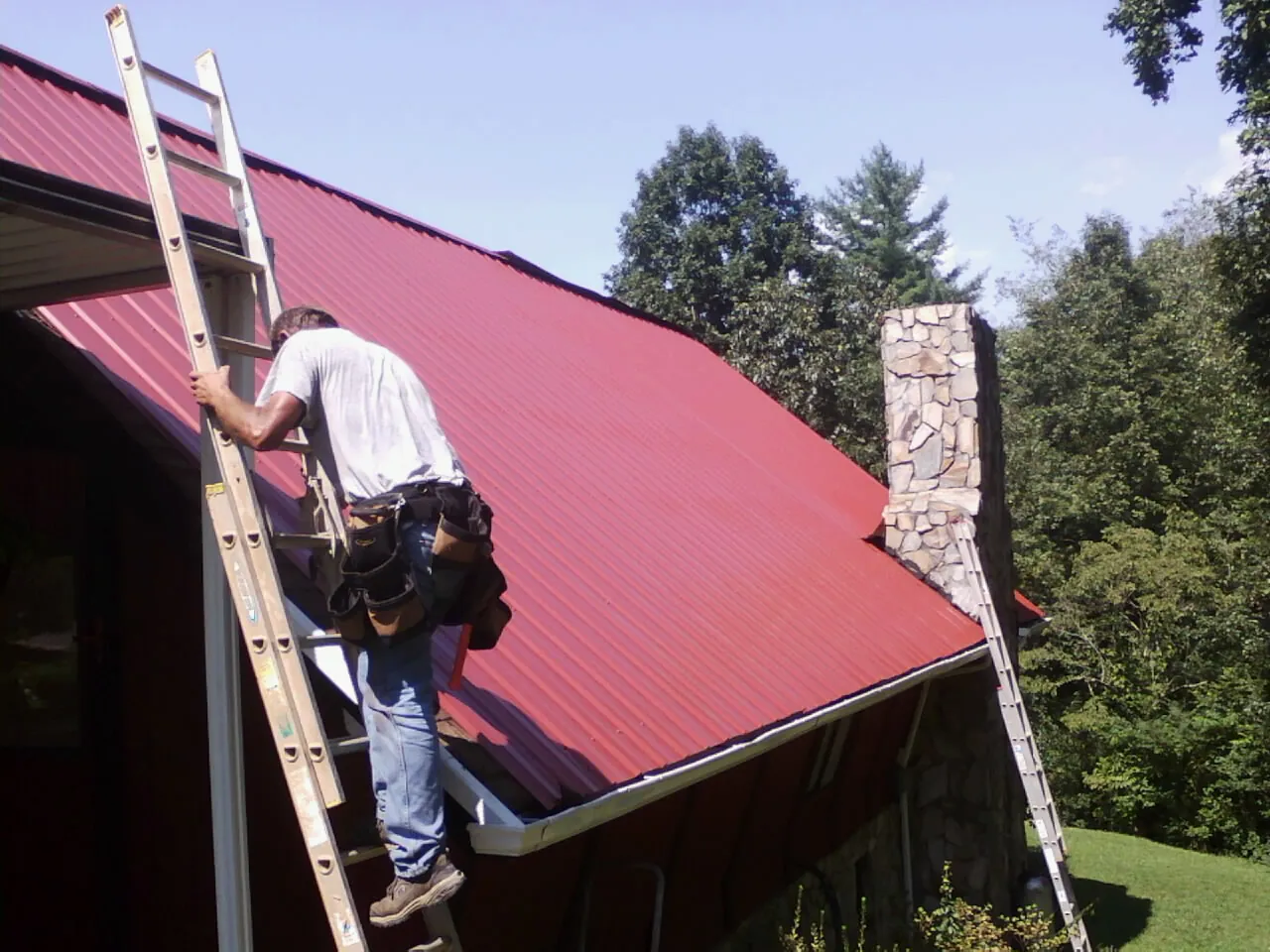 Roofer on a ladder beside a red metal roof and stone chimney — Soffit & Fascia Repair by Roofing Brothers in Centerville